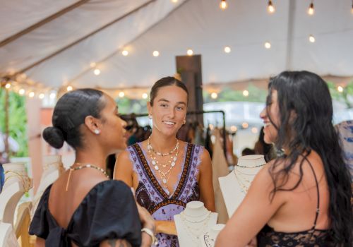 Women chat at an outdoor jewelry booth under warm string lights, with necklaces and bracelets displayed on stands.