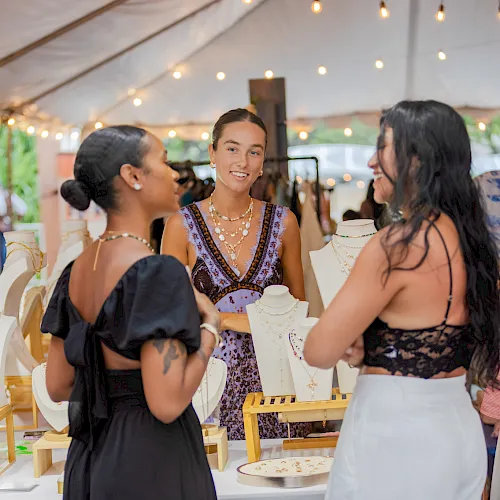Women chat at an outdoor jewelry booth under warm string lights, with necklaces and bracelets displayed on stands.