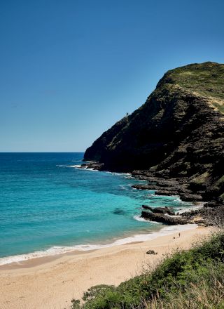 A sunny coastal scene with a sandy beach, turquoise waves, and a rocky cliff towering over the shore.