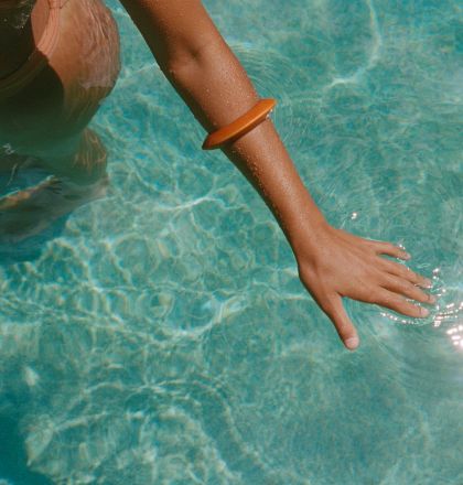 A person&rsquo;s hand reaching into a clear blue swimming pool, with sunlight glinting on the water&rsquo;s surface, creating a shimmering effect.