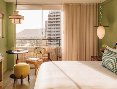 A bright, cozy hotel room with a large bed, light wood furniture, a desk, and a window view of a distant city skyline.