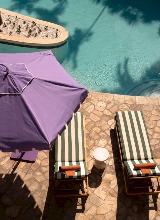 Two striped lounge chairs with towels under a purple patio umbrella beside a pool, on a stone deck with a small round table.