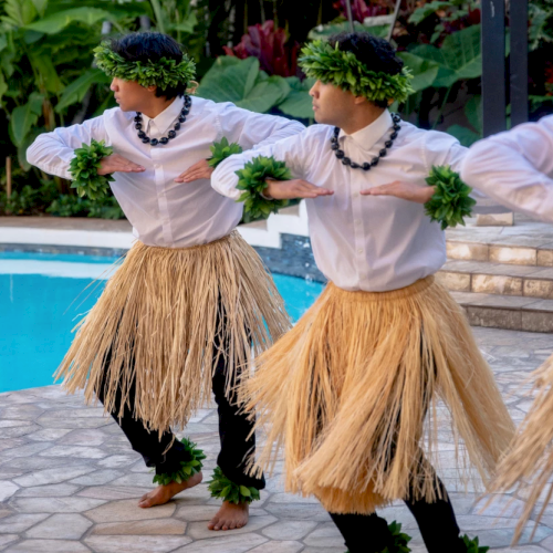 Three people perform a traditional Hawaiian dance by a pool, wearing grass skirts, leis, white shirts, and leafy headbands.