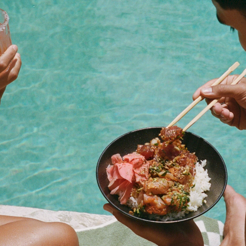 Two people by a pool enjoy a meal: a bowl of rice with meat/veg topped with sauce, while one sips a pink drink with a straw, all relaxed by blue water.