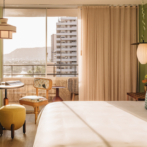Una acogedora habitaci&oacute;n de hotel con una pared decorativa verde, cama con almohadas a cuadros, un puf redondo amarillo, mesitas auxiliares, balc&oacute;n con vistas a los edificios de la ciudad y c&aacute;lida luz natural.