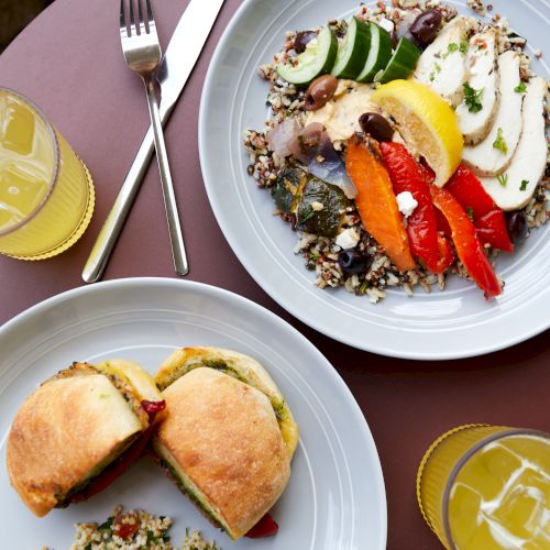 Un brunch abundante: cuencos repletos de verduras con pepinos, pimientos y c&iacute;tricos en rodajas en un plato, un s&aacute;ndwich sustancioso con verduras en el otro, adem&aacute;s de zumo de naranja.