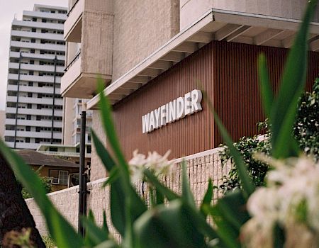 A city building entrance with a “WAYFINDER” sign, framed by green plants and tall high-rise apartments in the background.