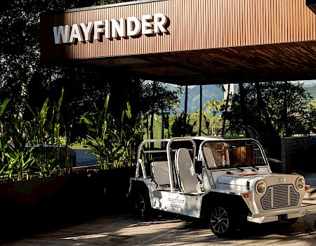 A white “Wayfinder” sign tops a wooden building, with a golf-cart style tour vehicle parked amid tropical plants.