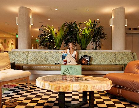 A woman lounges on a patterned sofa in a hotel lobby, surrounded by large potted plants and warm lighting.