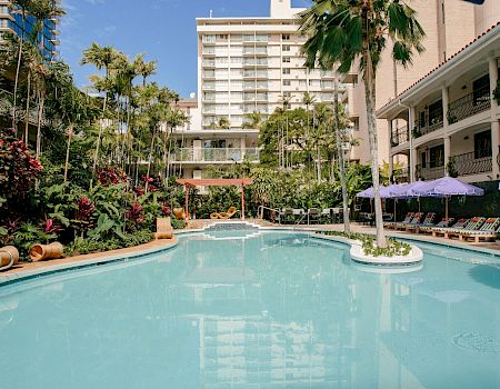 A sunny hotel courtyard with a large outdoor swimming pool, palm trees, lush gardens, and lounge chairs under purple umbrellas.