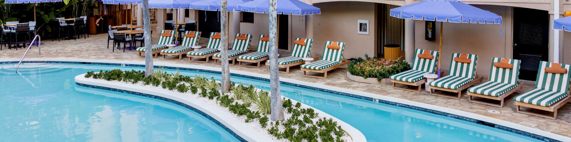 Tropical hotel courtyard with a bright pool, striped lounge chairs under blue umbrellas, and palm trees beside balcony rooms.
