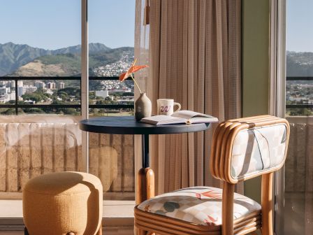 Sunlit balcony seating: wood chair and round table with open book, mug, vase, and hanging lamp beside a mountain view.