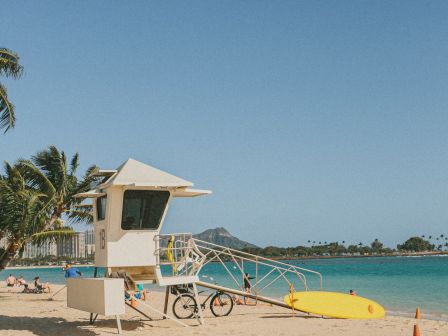 A lifeguard tower sits on a sunny sandy beach beside a yellow surfboard and orange cones under a clear blue sky.