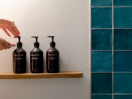Three Grown Alchemist pump bottles sit on a shelf as hands reach to dispense in a bathroom beside blue tile.