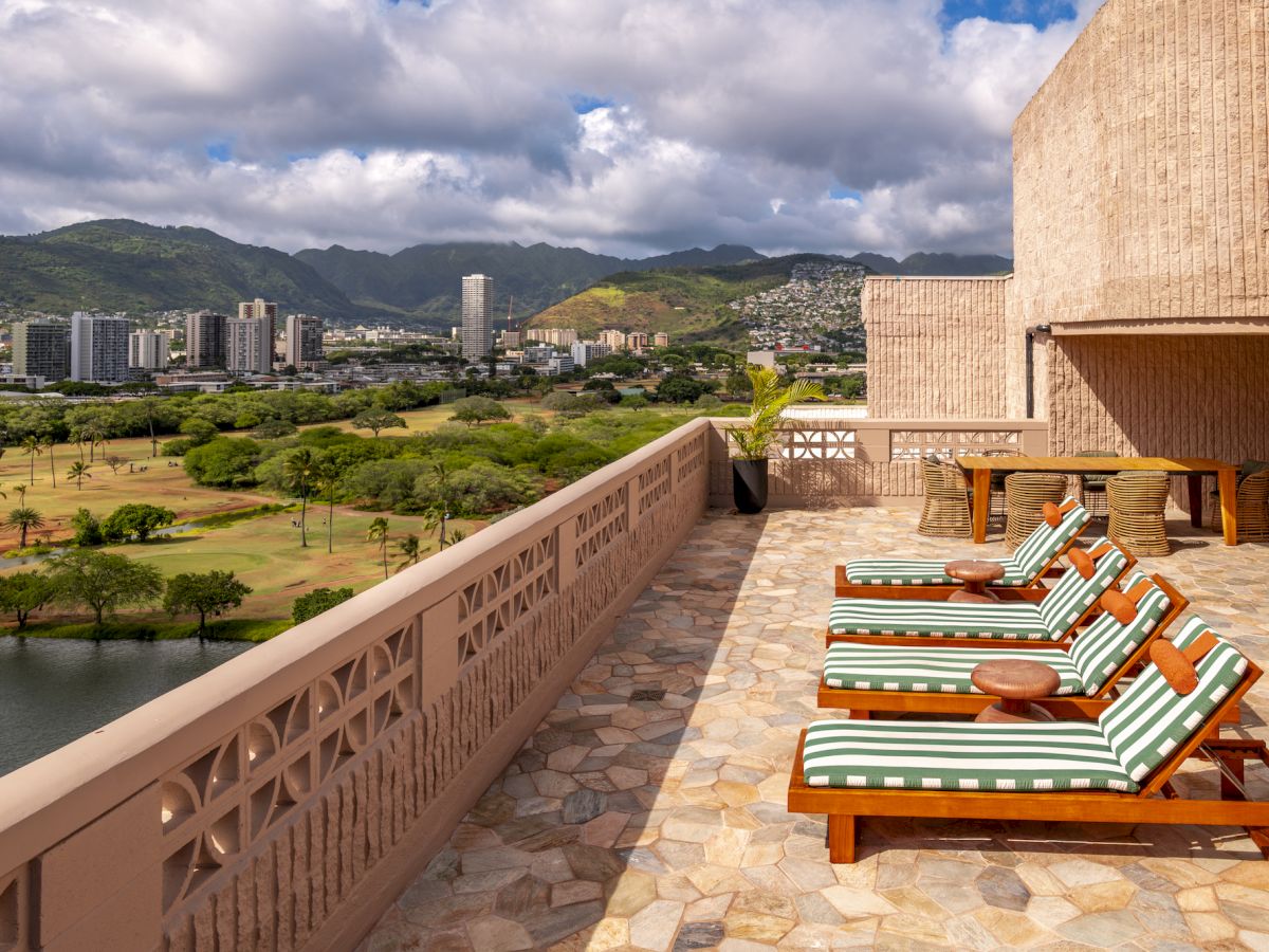 Balcon d'h&ocirc;tel avec chaises longues ray&eacute;es, donnant sur une vall&eacute;e verdoyante, un lac, des b&acirc;timents de la ville et des montagnes sous un ciel nuageux.