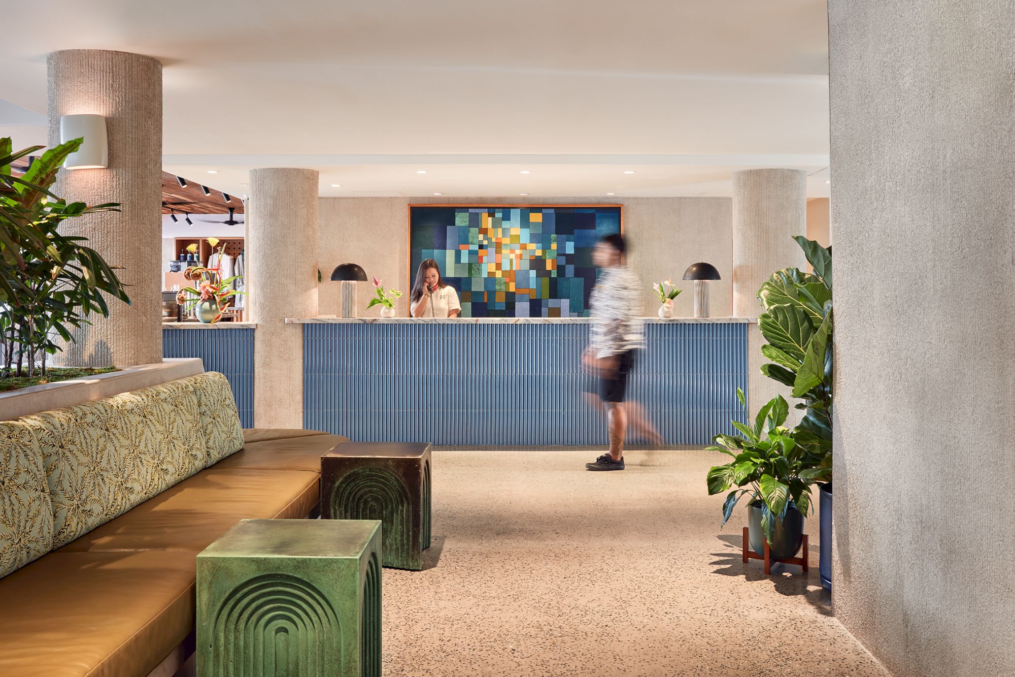 A modern hotel lobby with plants, a striped reception counter, colorful wall art, and people walking past.