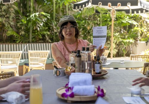 A woman in an outdoor setting holds a flyer for a hand massage & scrub workshop beside essential-oil bottles on a table.