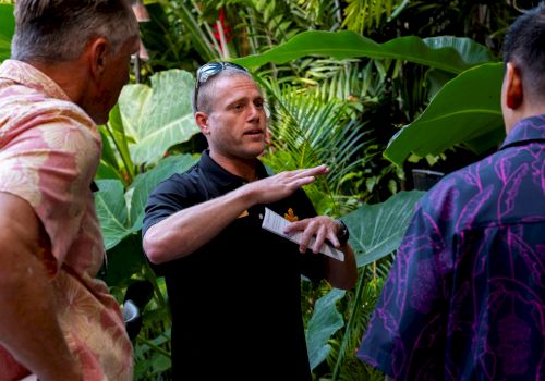 A tour guide gestures while explaining plants in a lush garden filled with large green leaves.