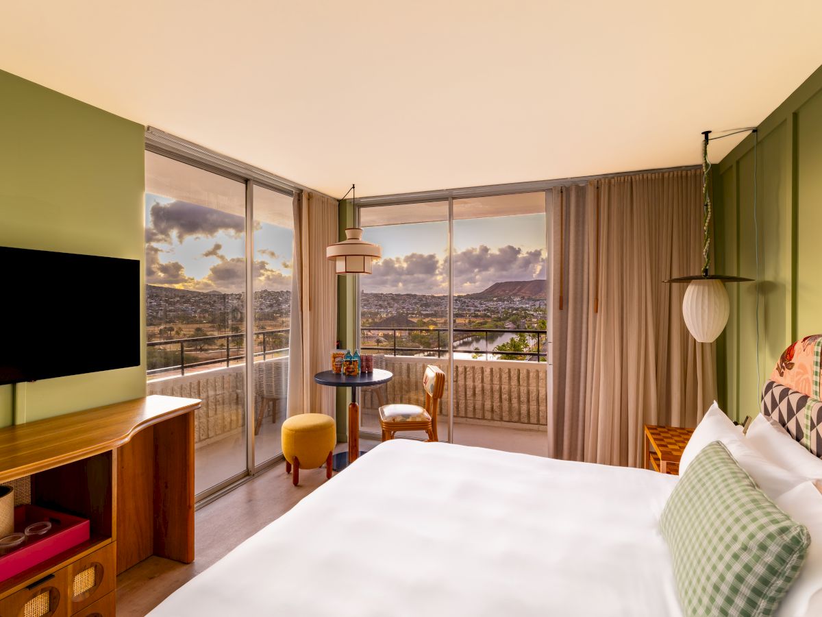 A stylish hotel room with a bed, TV, and balcony doors opening to a sunset city view over a palm-lined valley.