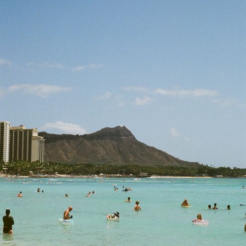 People float in a turquoise ocean at a sunny beach, with resorts and a dramatic mountain backdrop behind the water.