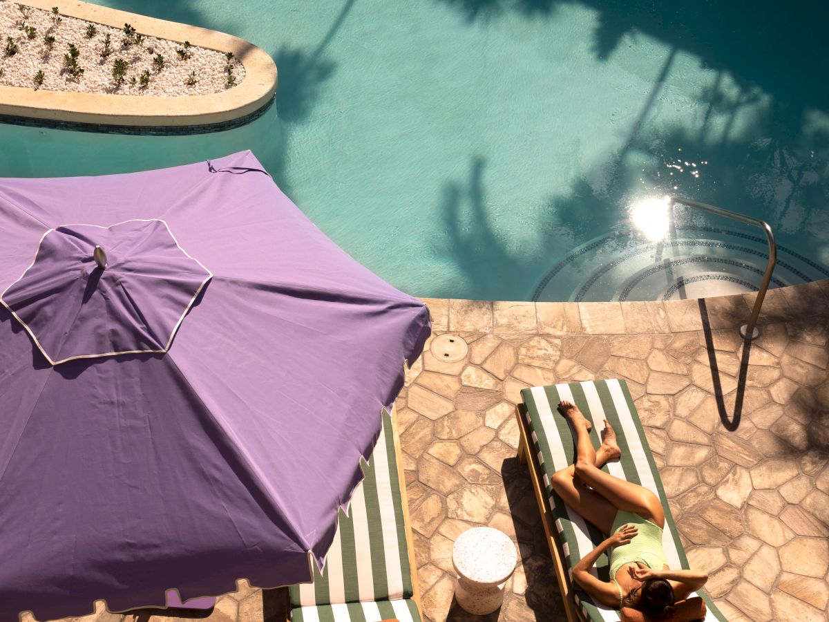 A poolside scene with turquoise water, lounge chairs, a purple umbrella, and a person relaxing in the sun.