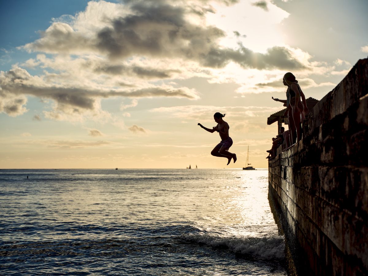 A group of kids and a woman leap into the ocean from a wooden dock at sunset, under dramatic clouds.