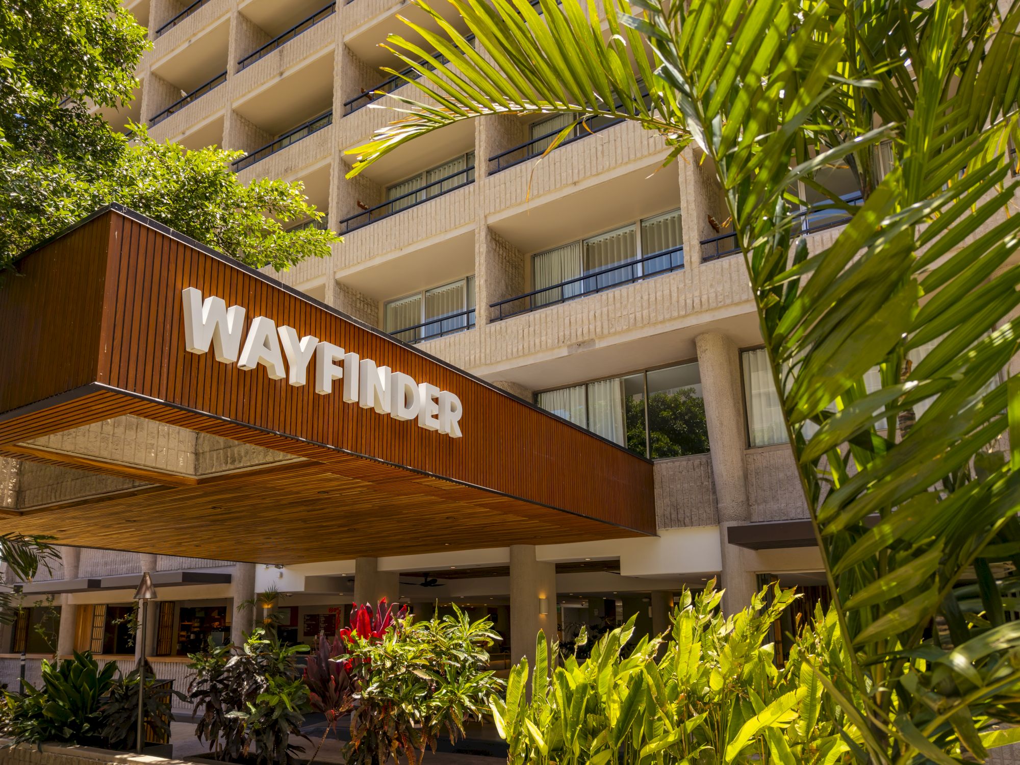 A resort courtyard with palm leaves, a multi-story hotel facade, and the word &ldquo;WANDER&rdquo; lit on a wooden wall.