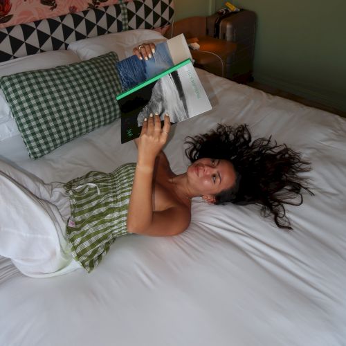 A person lounges in bed with a book, under a bedside lamp in a cozy room.