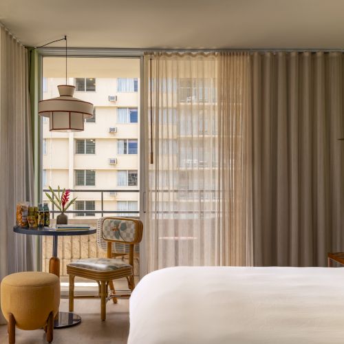 A bright hotel room with a bed, sheer curtains, a small chair and table, plus pendant lamps by the window.